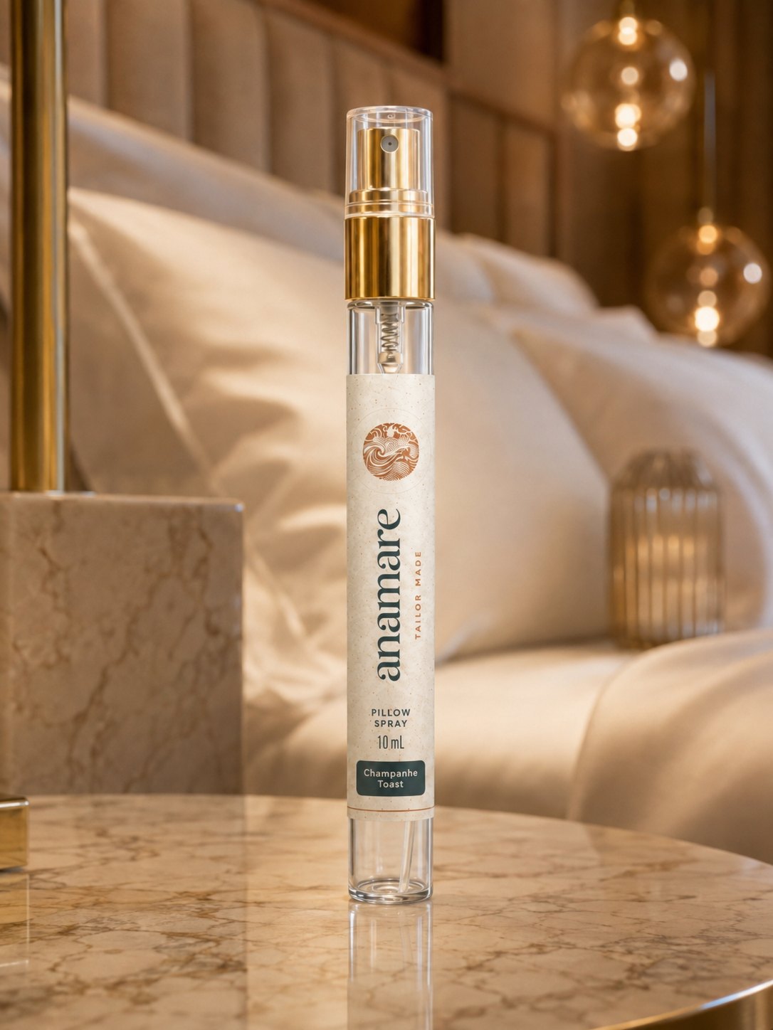 Pillow Spray Champanhe Toast 10 mL, flaconete de vidro com válvula spray dourada, em mesa de mármore dourado, com luminárias de vidro âmbar e roupa de cama acetinada ao fundo
