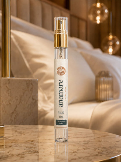 Pillow Spray Champanhe Toast 10 mL, flaconete de vidro com válvula spray dourada, em mesa de mármore dourado, com luminárias de vidro âmbar e roupa de cama acetinada ao fundo