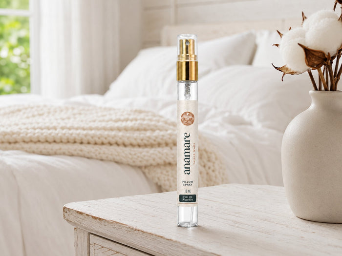 Pillow Spray Flor de Algodão 10 mL, flaconete de vidro com válvula spray dourada, em aparador de madeira patinada, com ramos de algodão em vaso de cerâmica, manta de tricô e cama branca ao fundo