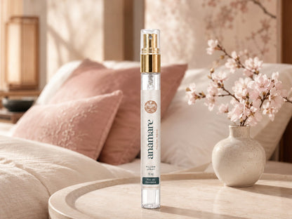 Pillow Spray Flor de Cerejeira 10 mL, flaconete de vidro com válvula spray dourada, em mesa redonda de mármore claro, com ramo de cerejeira em flor em vaso de cerâmica e almofadas rosadas ao fundo