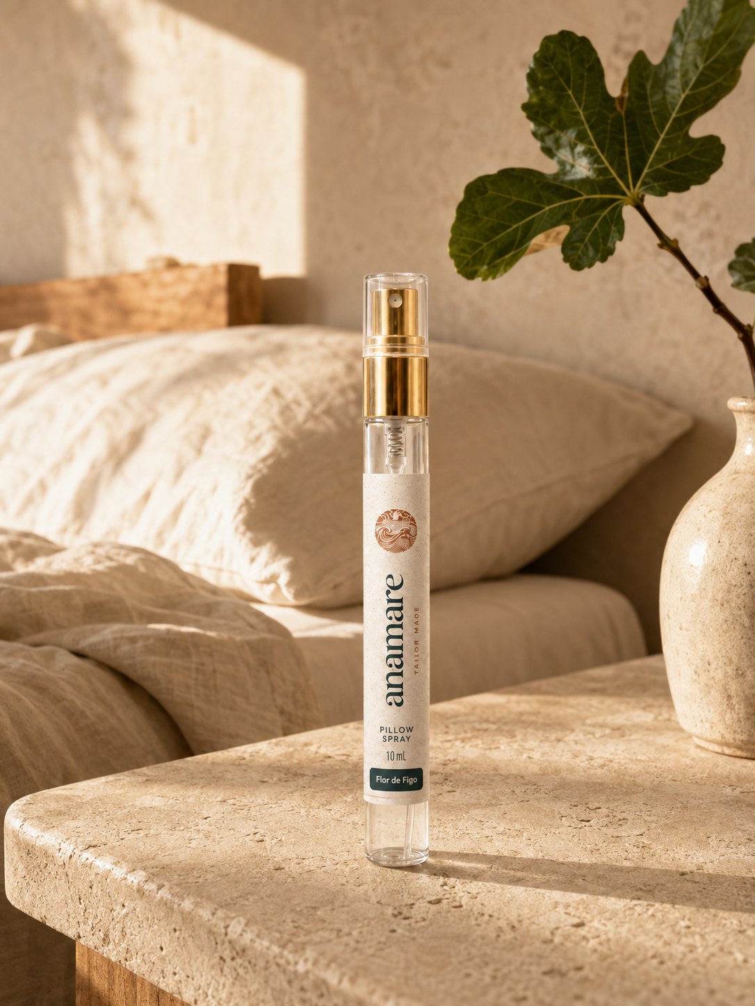 Pillow Spray Flor de Figo 10 mL, flaconete de vidro com válvula spray dourada, em mesa de travertino ao sol, com galho de figueira em vaso de cerâmica e almofadas de linho cru ao fundo