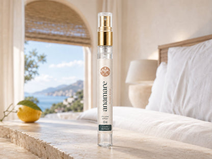 Pillow Spray Limão Siciliano 10 mL, flaconete de vidro com válvula spray dourada, em bancada de travertino diante de arco aberto para o mar, com limão-siciliano ao lado e cama de linho branco ao fundo