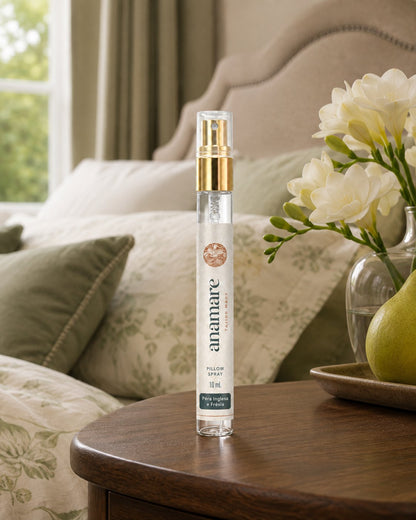 Pillow Spray Pera Inglesa e Frésia 10 mL, flaconete de vidro com válvula spray dourada, em mesa de cabeceira de madeira escura, com frésias brancas em jarro de vidro, pera em bandeja de madeira e cabeceira estofada ao fundo