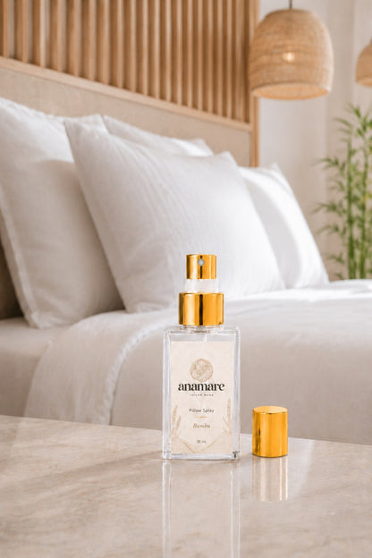 Pillow Spray Bambu 30 mL, frasco de vidro com válvula e tampa douradas, em quarto de ripado de madeira com luminárias de fibra natural e bambu ao fundo, sobre bancada de mármore bege