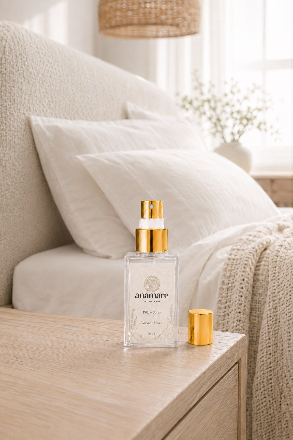 Pillow Spray Flor de Algodão 30 mL, frasco de vidro com válvula e tampa douradas, em quarto claro com cabeceira de bouclé, roupa de cama branca e manta de tricô, sobre criado-mudo de carvalho