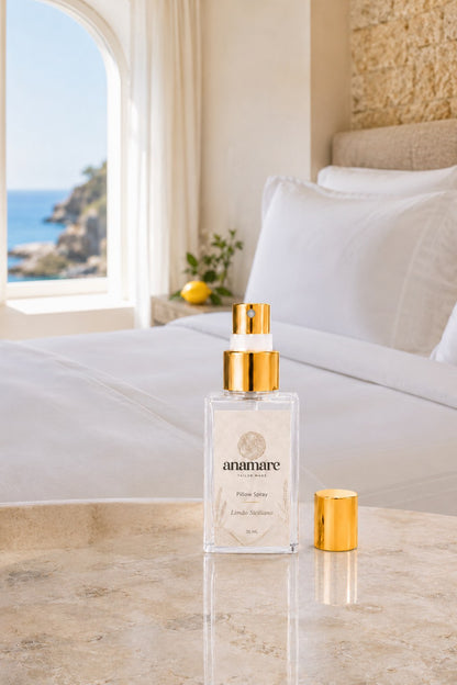 Pillow Spray Limão Siciliano 30 mL, frasco de vidro com válvula e tampa douradas, em quarto mediterrâneo com janela em arco para o mar, parede de pedra e limões frescos, sobre bancada de mármore claro