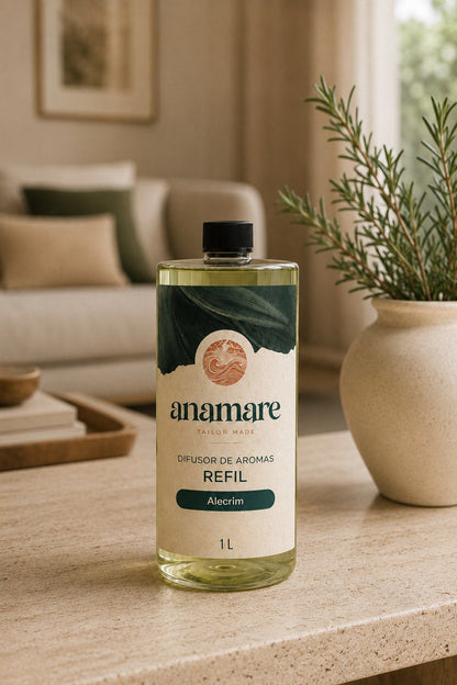 Refil de Difusor de Aromas Anamare de 1 litro, frasco transparente com tampa preta e rótulo verde-escuro com o selo da marca, sobre mesa de travertino em sala de estar, com sofá de linho, almofadas verdes e alecrim em vaso de cerâmica ao fundo