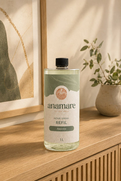Refil de Home Spray Anamare de 1 litro, frasco transparente com tampa preta e rótulo verde-sálvia com o selo da marca, sobre aparador de madeira clara ripada, com quadro de arte abstrata e vaso de cerâmica ao fundo em luz natural