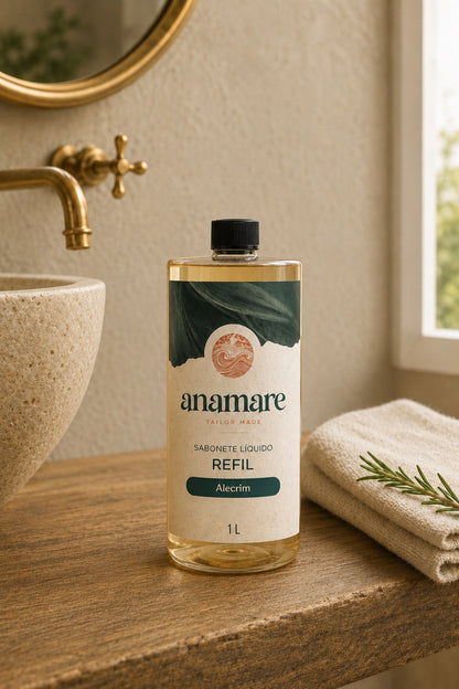 Refil de Sabonete Líquido Anamare de 1 litro, frasco transparente com tampa preta e rótulo de papel com faixa verde, sobre bancada de madeira em lavabo com cuba de pedra, torneira dourada, espelho redondo e toalha de linho com ramo de alecrim