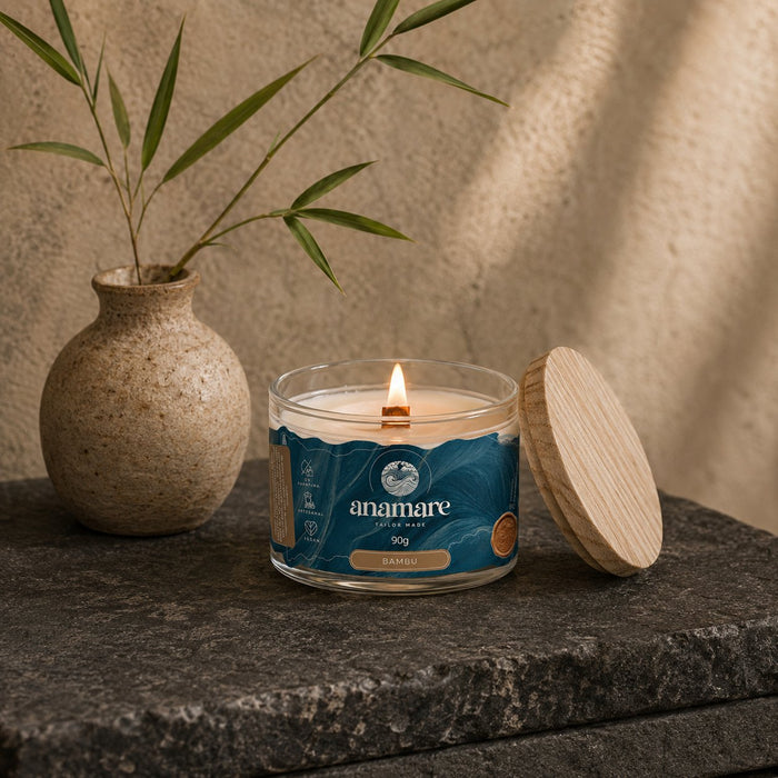 Vela Aromática Bambu Moyen 90 g acesa, pote de vidro com pavio de madeira, em bancada de pedra escura, com hastes de bambu em vaso de cerâmica, persiana de madeira e tampa de madeira ao lado