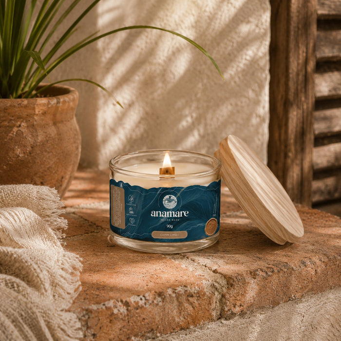 Vela Aromática Capim Limão Moyen 90 g acesa, pote de vidro com pavio de madeira, em bancada de pedra, com capim verde em vaso de barro, manta de linho franjada e tampa de madeira ao lado