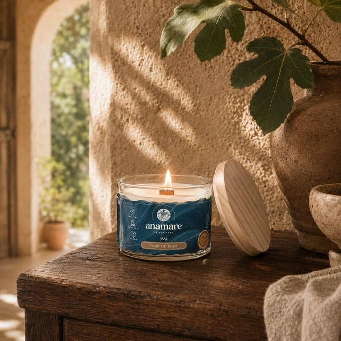 Vela Aromática Flor de Figo Moyen 90 g acesa, pote de vidro com pavio de madeira, em aparador de madeira rústica sob arco de pedra, com folhas de figueira, vaso de barro e tampa de madeira ao lado