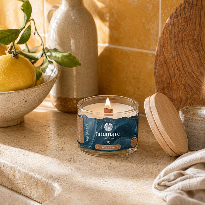Vela Aromática Limão Siciliano Moyen 90 g acesa, pote de vidro com pavio de madeira, em bancada de pedra ao sol, com limões-sicilianos em tigela de cerâmica, azulejos amarelos ao fundo e tampa de madeira ao lado