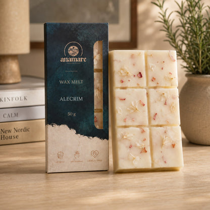 Wax Melt Anamare Alecrim de 50 g, barra de cera vegetal com pétalas secas ao lado da caixa azul-petróleo com janela, em escrivaninha de madeira clara com livros empilhados e alecrim fresco em vaso de cerâmica