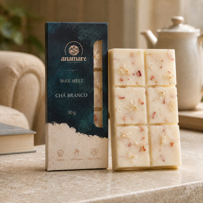 Wax Melt Anamare Chá Branco de 50 g, barra de cera vegetal com pétalas secas ao lado da caixa azul-petróleo com janela, em canto de leitura com poltrona clara, livro fechado e bule de porcelana sobre bancada de mármore bege