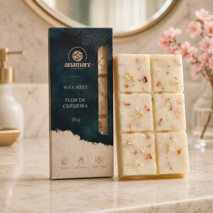 Wax Melt Anamare Flor de Cerejeira de 50 g, barra de cera vegetal com pétalas secas ao lado da caixa azul-petróleo com janela, em lavabo de mármore com espelho redondo dourado, saboneteira de porcelana e galho de cerejeira em flor