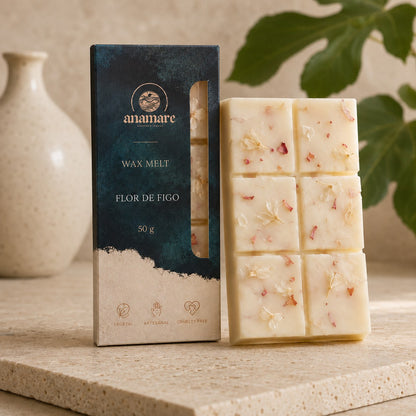 Wax Melt Anamare Flor de Figo de 50 g, barra de cera vegetal com pétalas secas ao lado da caixa azul-petróleo com janela, em bancada de travertino com vaso de cerâmica clara e folha de figueira ao fundo